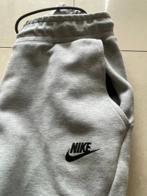 Nike Tech Fleece sweatpants  - Snygga grå mjukisbyxor från Nike med svart logga på låret. Byxorna har dragsko i midjan, ribbade muddar vid bensluten och praktiska sidofickor. Perfekta för en avslappnad och sportig stil. Tillverkade i mjukt och bekvämt material.