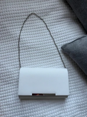 Vit kuvertväska med kedja - Snygg vit kuvertväska med stilren silverfärgad kedja och metallspänne. Väskan har en slät, lätt strukturerad yta och en klassisk rektangulär form. Perfekt för dig som vill ha en enkel men elegant accessoar till din outfit. Endast använd en gång och är perfekt till balen! ❤️