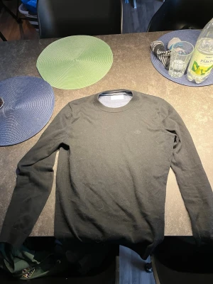 Svart stickad tröja från Tiger of Sweden - Snygg svart stickad tröja från Tiger of Sweden med rund halsringning och diskret broderad logga på bröstet. Tröjan har långa ärmar och ribbade muddar vid hals, ärmslut och nederkant. Perfekt basplagg till garderoben.