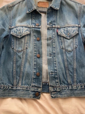 Klassisk blå Levi's jeansjacka - Storlek S. Säljer en klassisk blå jeansjacka från Levi's med snygg tvätt och bruna metallknappar. Jackan har två bröstfickor med lock och knapp samt den ikoniska röda Levi's-taggen. Perfekt passform och tidlös stil som funkar till allt. 