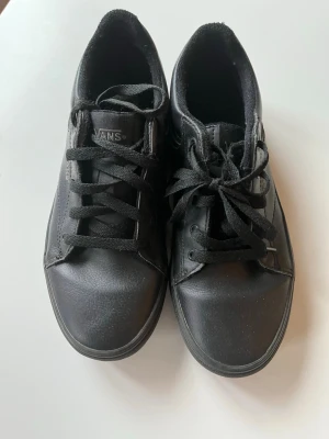 Svarta Vans sneakers i läder - Säljer ett par helsvarta Vans sneakers i lågt modell med klassisk design. Skorna är tillverkade i läder med matchande svarta skosnören och diskret Vans-logga på sidan. Perfekta för dig som gillar en clean och stilren look.
