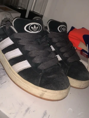 Adidas Campus svarta sneakers - Svarta Adidas Campus sneakers med klassiska vita ränder på sidorna och stor vit logga på plösen. Ovandelen är i mocka och sulan är vit med gummidetaljer. Skorna har breda svarta skosnören och en tidlös streetstil som passar perfekt till jeans eller joggers. och man kan få snygga tjejer med dem