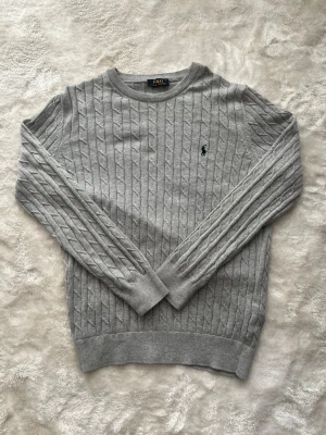 Ljusgrå kabelstickad tröja Polo Ralph Lauren - Snygg ljusgrå kabelstickad tröja från Polo Ralph Lauren med rund halsringning och den klassiska broderade loggan på bröstet. Tröjan har ribbade muddar vid ärmslut och nederkant och är tillverkad i ett mjukt material som passar perfekt till alla tillfällen.