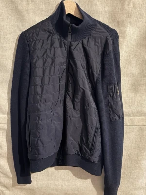 Marinblå cardiganjacka från J.Lindeberg - Snygg marinblå cardiganjacka från J.Lindeberg med  stickade ärmar. Jackan har hög krage, dragkedja framtill och en praktisk ficka med logga på ärmen. Perfekt för lager-på-lager och stilrena outfits.