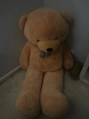 Stor beige nallebjörn med rosett - Jättestor mjuk nallebjörn i beige plysch med rund nos och svart-vit rutmönstrad rosett runt halsen. Perfekt som mysig inredningsdetalj eller present. Sitter lutad mot väggen och har långa ben och stora tassar. Jätte stor o mysig