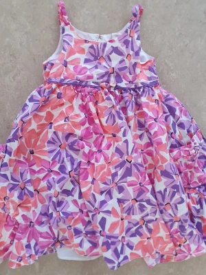 Klänning - Söt blommig klänning från Maggie & Zoe i vitt, rosa, lila och orange. Klänningen har smala axelband och ett utställt, luftigt fall. Yttertyget är i bomull och fodret i polyester och bomull. Perfekt för varma dagar!