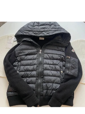 Moncler Cardigan  - Snygg svart cardigan från Moncler med stickade ärmar och huva. Jackan har quiltad front och rygg i glansigt material, dragkedja framtill och Moncler-logga på ärmen. Perfekt för kalla dagar och riktigt trendig look.