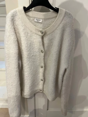 Vit fluffig kofta från Kappahl - Mysig vit kofta från Kappahl i storlek 158/164. Koftan är fluffig och har rund halsringning samt silverfärgade knappar framtill. Perfekt för lager på lager och enkel att matcha med olika outfits.