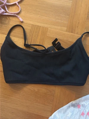 Svart ribbad bikiniöverdel - Snygg svart bikiniöverdel med ribbad struktur och raka axelband. Modellen har en enkel och stilren design med spänne i ryggen och justerbara band. Perfekt för strand eller poolhäng. Passar S. 