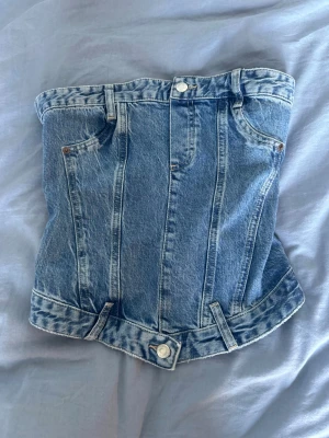 Denim korsett-topp med dragkedja - Cool korsett-topp i blå denim med klassiska jeansdetaljer som fickor, bälteshällor och knappar. Toppen har en markerad midja och dragkedja bak för enkel på- och avtagning. Perfekt för dig som vill sticka ut med en edgy look.