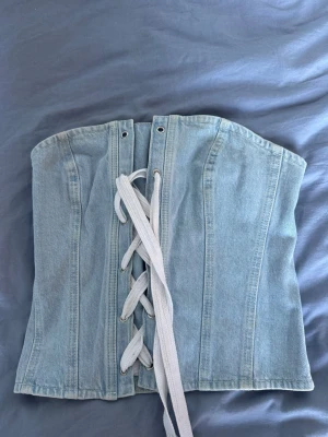 Ljusblå denimkorsett med snörning - Trendig ljusblå korsett i denim med vit snörning framtill. Modellen har markerade sömmar och metallöljetter för snörningen. Perfekt för dig som vill ha en edgy och modern look. Materialet är klassiskt jeansigt och ger en cool vibe.