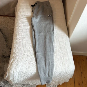 Gråa mjukisbyxor från Colourwear - Säljer ett par gråa mjukisbyxor med resår i midjan och svarta dragsnören. Byxorna har muddar vid bensluten och en liten logga på vänster lår. Perfekta för chill dagar eller när du vill vara bekväm. Materialet känns mjukt och stretchigt.