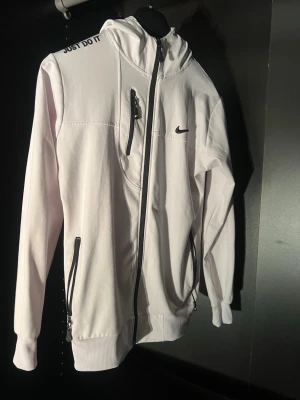 Vit Nike hoodie med dragkedja - Snygg vit hoodie från Nike med svart logga på bröstet och texten 'Just Do It' på axeln. Hoodien har dragkedja framtill, tre fickor med dragkedja och ribbade muddar. Tillverkad i mjukt material och har en huva med snörning.