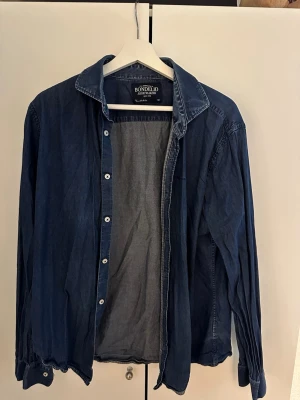Mörkblå jeansskjorta Bondelid - Snygg mörkblå jeansskjorta från Bondelid med klassisk krage och vita knappar. Skjortan har lång ärm och är tillverkad i mjuk bomull, perfekt för en avslappnad stil. Det står storlek XL men det stämmer inte, jag skulle säga att den passar M numera.