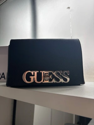 Svart kuvertväska från Guess - Snygg svart handväska från Guess i stilren design med stort silverfärgat logotypspänne framtill. Väskan är tillverkad i ett strukturerat konstläder och har en kompakt, rektangulär form. Perfekt accessoar för att lyfta din outfit. Jättefin skick framsidan är lite använd men syns inte mycket. Band tillkommer 