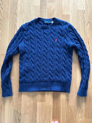 Marinblå kabelstickad tröja Polo Ralph Lauren - Kabelstickad marinblå tröja från Polo Ralph Lauren med rund halsringning och ribbade muddar. Tröjan har det klassiska röda Polo-logotypen broderad på bröstet. Perfekt för dig som gillar tidlös och stilren design.