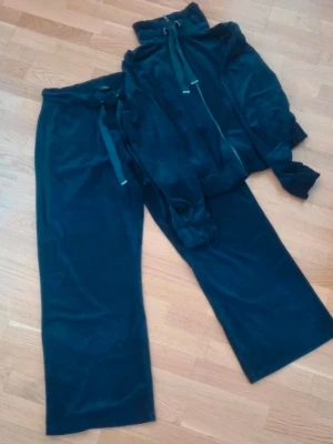 Svart velourset med dragkedja från Twilfit - Mysigt loungewear-set i svart velour med matchande byxor och ziptröja. Tröjan har dragkedja, hög krage och snörning i midjan. Byxorna har resår och snörning i midjan för skön passform. Perfekt för chill dagar hemma.