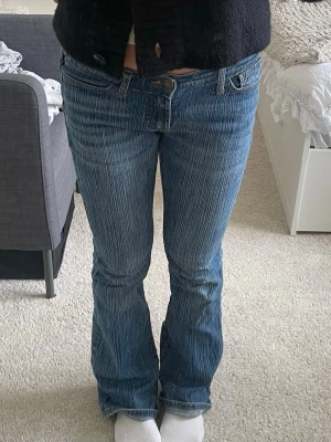 Blå bootcut jeans med låg midja - Säljer ett par blå bootcut jeans med snygga slitningar och ljusa tvättade detaljer. Jeansen har låg midja, klassiska fem fickor och dragkedja med knapp. Perfekta för en avslappnad och trendig look.