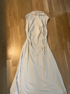 Lång ljus klänning med hög krage - Elegant långklänning i ljus beige ton med hög krage och ärmlös design. Klänningen har en figurnära passform upptill och faller mjukt nedåt. Materialet ser ut att vara satin eller polyester för en lyxig känsla. Perfekt för speciella tillfällen.