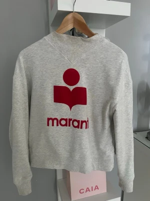 Grå sweatshirt från Isabel Marant - Säljer en ljusgrå sweatshirt från Isabel Marant med det ikoniska röda logotrycket på bröstet. Tröjan har rund halsringning, ribbade muddar och är gjord i mjukt bomullsmaterial. Perfekt för en avslappnad och trendig look.