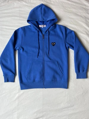 Blå hoodie från Comme des Garçons Play - Snygg blå hoodie med dragkedja från Comme des Garçons Play. Har en svart hjärtpatch med ögon på bröstet, två fickor framtill och justerbar huva med snören. Tillverkad i mjuk bomull för skön känsla och avslappnad stil.