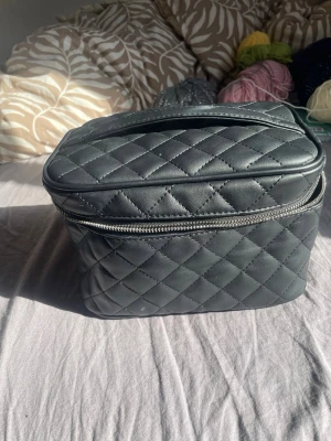 Svart quiltad necessärväska - Snygg svart quiltad necessärväska i konstläder med dragkedja runt om och ett praktiskt handtag upptill. Perfekt för att förvara smink, hudvård eller andra småprylar. Klassisk design med rutmönster som ger en lyxig känsla.