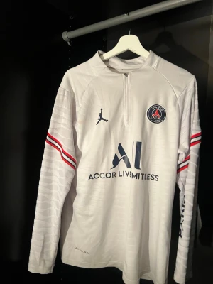 Vit PSG träningströja Jordan - Snygg vit långärmad träningströja från PSG och Jordan med halv dragkedja. Tröjan har röda och blå ränder på ärmarna, PSG-logga på bröstet och tryck som 'Accor Live Limitless' och 'Visit Rwanda'. Perfekt för fotbollsträning eller chill. Tillverkad i lätt funktionsmaterial. Sitter vanligt på S , Sitter slim fit på M - L. 