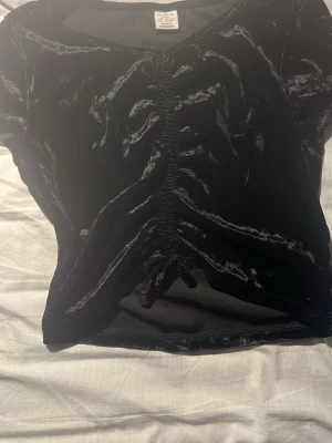 Svart sammets topp med snörning - Snygg svart topp från Lindex  i mjuk sammet med korta ärmar och snörning framtill som ger en cool rynkad effekt. Perfekt för dig som vill ha en trendig och unik look. Toppen har en lätt croppad passform och känns riktigt skön mot huden.