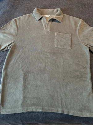 Olivgrön frotté polotopp från East West - Säljer en olivgrön polotopp från East West i mjuk frotté. Toppen har korta ärmar, klassisk krage och en bröstficka. Perfekt för en chill och avslappnad stil.