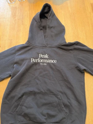 Svart hoodie Peak Performance - Säljer en svart hoodie från Peak Performance med stor huva och magficka. Klassisk logga broderad i vitt på bröstet och mjukt material som känns skönt mot huden.