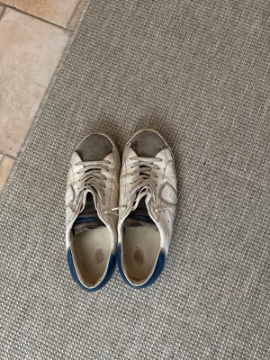 Philippe model skor - Säljer ett par klassiska vita sneakers med blå detalj på hälen och grå inslag på tån. Skorna har en låg profil, snörning och är tillverkade i läder med textilfoder. Perfekt för dig som gillar en clean och enkel stil.