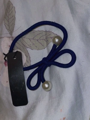 Blå hårsnodd med pärldetaljer - Snygg blå hårsnodd i elastiskt material med dekorativ rosett och två vita pärlliknande detaljer i ändarna. Perfekt för att piffa till din frisyr med en söt accessoar.