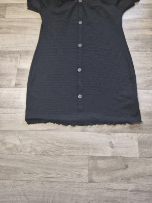 Svart kortklänning med knappar - En svart kortklänning med knappar framtill och korta ärmar. Klänningen har en lätt rynkad struktur och volangkant vid halsringning och nederkant. Perfekt för dig som gillar enkel men snygg stil. 4 xl