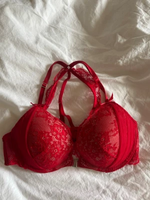 Röd spets-bh med dubbla axelband - Snygg röd bh med vadderade kupor och dekorativa dubbla axelband. Bh:n är täckt av blommig spets och har en liten rosett och smycke framtill. Justerbara band för bästa passform. Bh:n är från hunkemöller, använd få gånger