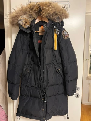 Svart dunjacka från Parajumpers - Lång svart dunjacka från Parajumpers med stor huva och avtagbar pälsdetalj. Jackan har dragkedja, knappar, flera fickor och en gul detalj vid halsen. Perfekt för kalla vinterdagar och riktigt snygg streetstil.