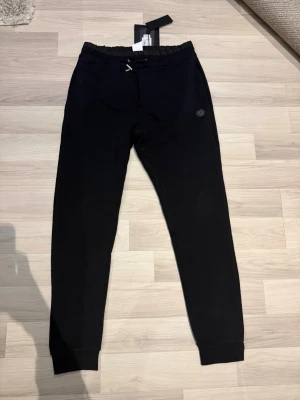 Svarta mjukisbyxor från Philipp Plein - Snygga svarta mjukisbyxor från Philipp Plein med elastisk midja och ribbade benslut. Byxorna har en diskret logga på benet och dragkedja vid fickan. Helt nya, aldrig använt, kostade 4 tusen 