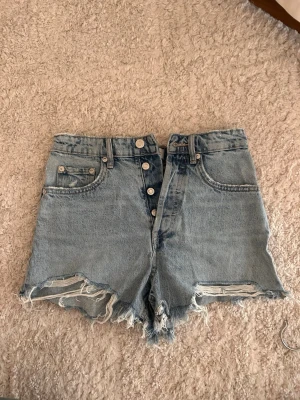 Högmidjade ljusblå jeansshorts - Säljer ett par högmidjade ljusblå jeansshorts med råa, fransiga kanter och slitningar framtill. Shortsen har knappar i gylfen och klassiska fem fickor. Perfekta för sommaren och ger en avslappnad look.