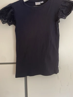 Mörkblå topp med spetsärmar Gina Tricot - Supersnygg mörkblå topp från Gina Tricot med korta ärmar i spets och broderade detaljer. Toppen är ribbad i materialet och har en normal passform. Perfekt för dig som vill ha något enkelt men ändå med en twist.