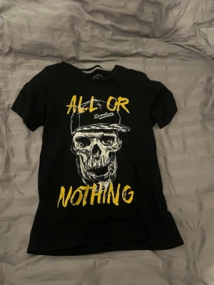 Svart t-shirt med gul text - Snygg svart t-shirt med trycket 'ALL OR NOTHING' i gul text och motiv på framsidan. T-shirten har rund hals och korta ärmar, perfekt för en avslappnad streetwear-look. Materialet känns mjukt och skönt mot huden.