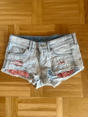 Lågmidjade ljusblåa denimshorts med slitningar  - Säljer ett par ljusblå denimshorts från HM med slitningar framtill. Köpte på Vinted men passade tyvärr inte mig. Under slitningarna syns de röda fickorna med vitt mönster. Klassiska fickor och knappgylf. Perfekta för sommaren!
