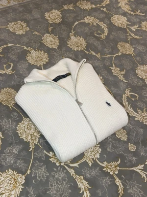 Vit stickad zip-tröja från Polo Ralph Lauren - Säljer en vit stickad tröja från Polo Ralph Lauren med silverfärgad dragkedja och klassisk broderad logga på bröstet. Tröjan har ribbad struktur och hög krage, perfekt för att hålla värmen. Enkel och stilren design som passar till många outfits.