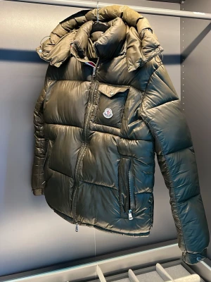 Olivgrön dunjacka från Moncler - Snygg olivgrön dunjacka från Moncler med hög krage, stor huva och dragkedja framtill. Jackan har en bröstficka med lock och Moncler-logga, samt två sidofickor med dragkedja. Puffig modell i glansigt material. Storlek M men passar även S