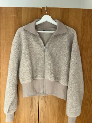 Beige teddyjacka med dragkedja - Mysig beige teddyjacka med hög krage och silverfärgad dragkedja framtill. Jackan har breda muddar i ärmslut och midja samt en croppad passform. 