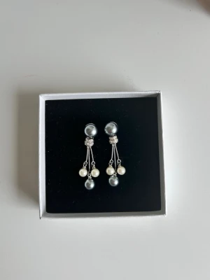 Silverfärgade örhängen med pärlor - Nya örhängen som aldrig har varit använda. Snygga örhängen i silverfärg med både vita och grå pärlor. Örhängena har tre hängande pärlor på varje sida och dekoreras med små gnistrande detaljer vid fästet. Perfekta för dig som gillar en elegant och trendig look.