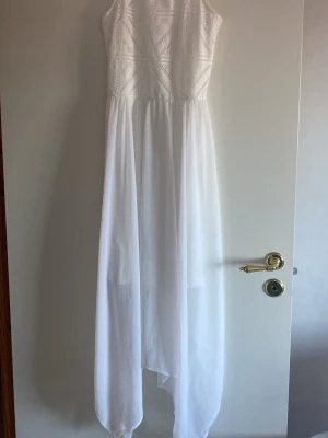 Vit långklänning  - Elegant vit långklänning med spetsmönstrad överdel och flowy kjol i tunt tyg. Klänningen har ärmlös design och rund halsringning, perfekt för sommarens speciella tillfällen. Stilren och feminin look.