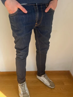Mörkblå Nudie Jeans  - Säljer ett par mörkblå slim fit jeans från Nudie Jeans med snygga slitningar och kontrastsömmar. Jeansen har klassisk femficksmodell, orange broderi på bakfickorna och är tillverkade i stretchig denim för skön passform. Storleken står nedan. Perfekta nu till våren. Hör av er vid frågor! 