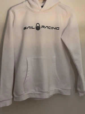 Vit hoodie från Sail Racing - Säljer en vit hoodie från Sail Racing med svart logga tryckt på bröstet. Hoodien har en klassisk känguruficka framtill och en skön huva. Perfekt för dig som gillar enkel och stilren design. Materialet känns mjukt och bekvämt.