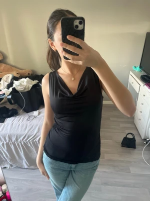 Svart ärmlös linnetopp - Snygg svart ärmlös linnetopp med lätt drapering vid halsen. Toppen har en figurnära passform och är perfekt att styla med jeans eller kjol. Materialet känns mjukt och stretchigt, vilket gör den bekväm att bära hela dagen.