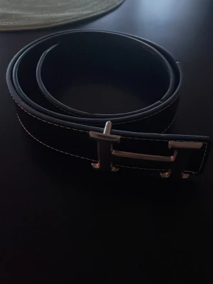 Svart skärp med silverfärgat H-spänne - Snyggt svart bälte med kontrastsömmar och ett stort H-format spänne i silverfärg. Bältet har en klassisk design och passar perfekt till jeans eller kostymbyxor. Tillverkat i skinn för en stilren och tidlös look.