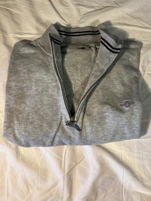 Grå halvzip-tröja från GANT - Snygg grå stickad halvzip-tröja från GANT med ribbad krage och svarta ränder. Tröjan har en liten broderad logga på bröstet och är tillverkad i bomull. Perfekt för en stilren och avslappnad look. Sparsamt andvänd